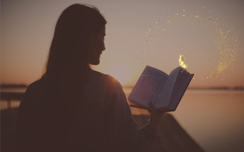 Eine Autorin liest am Strand in ihrem neu veröffentlichten Buch. Ein goldener Schmetterling zeigt den Zauber, der ein eigenes Buch umgibt.